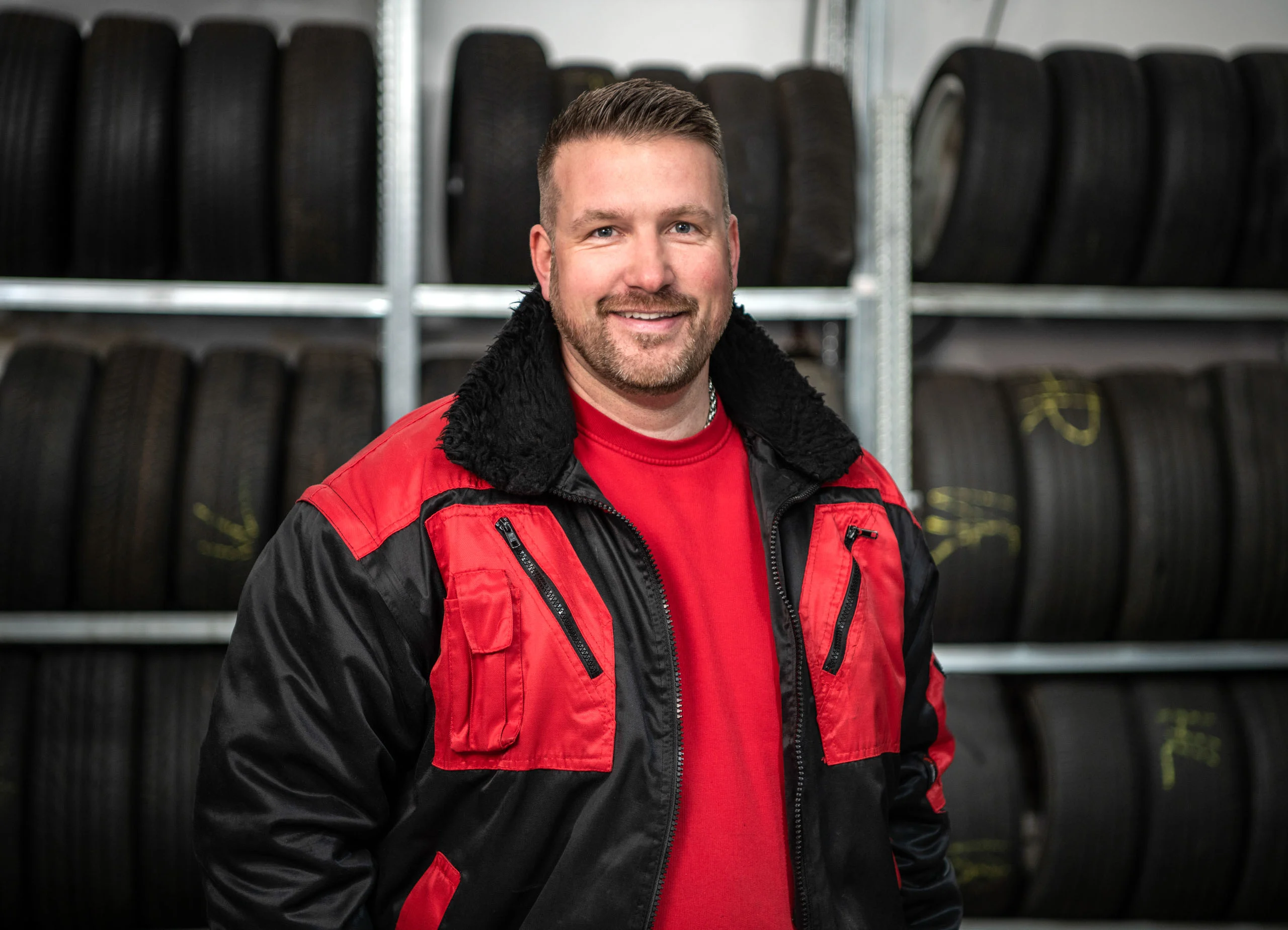 Business Portraits Fotogalerie 027 | Peter Lauritis Photography Kfz-Mechaniker in rot-schwarzer Arbeitsjacke vor einem Reifenregal