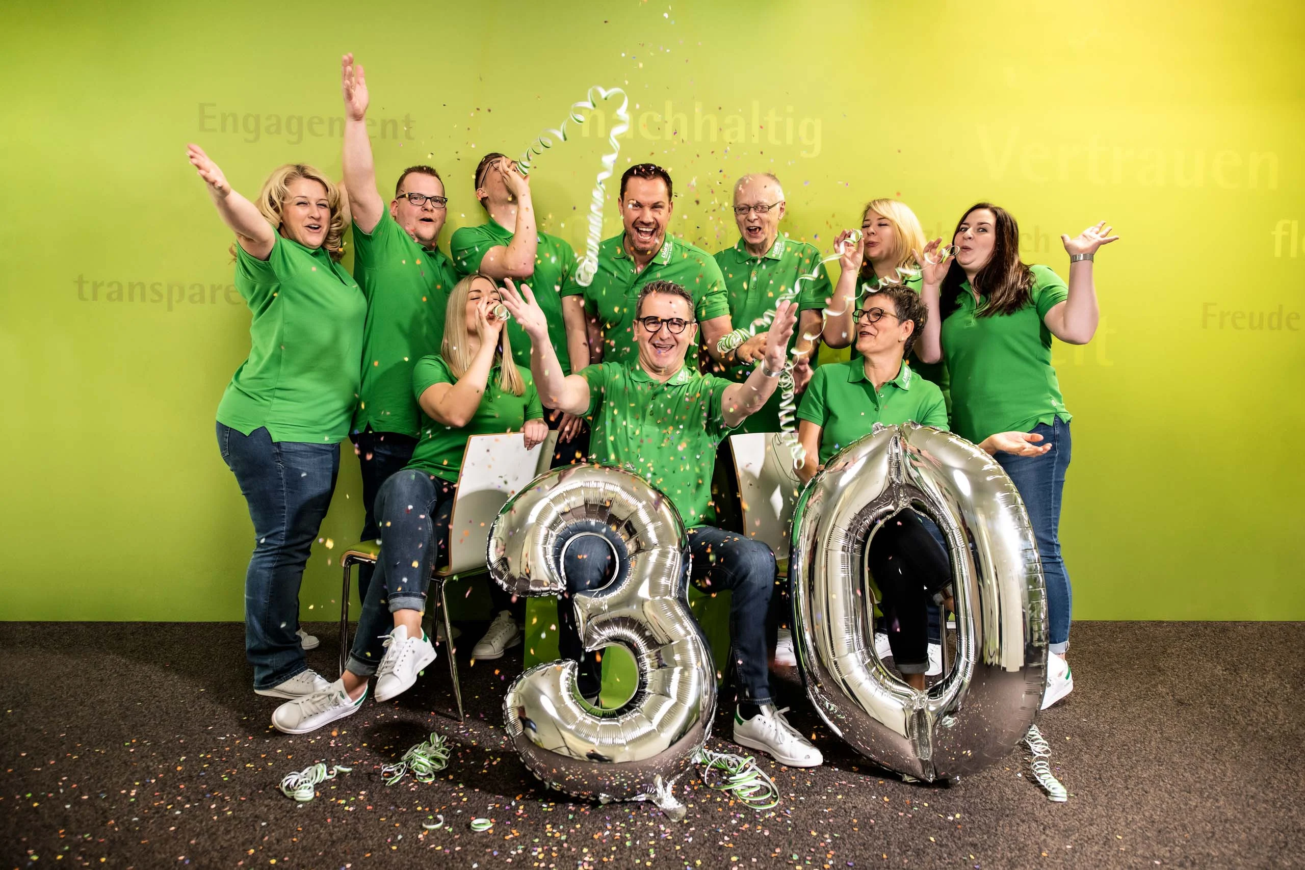 Business Portraits Fotogalerie 033 | Peter Lauritis Photography Team in Grün jubelt mit Ballons „30“ und Konfetti vor grüner Wand