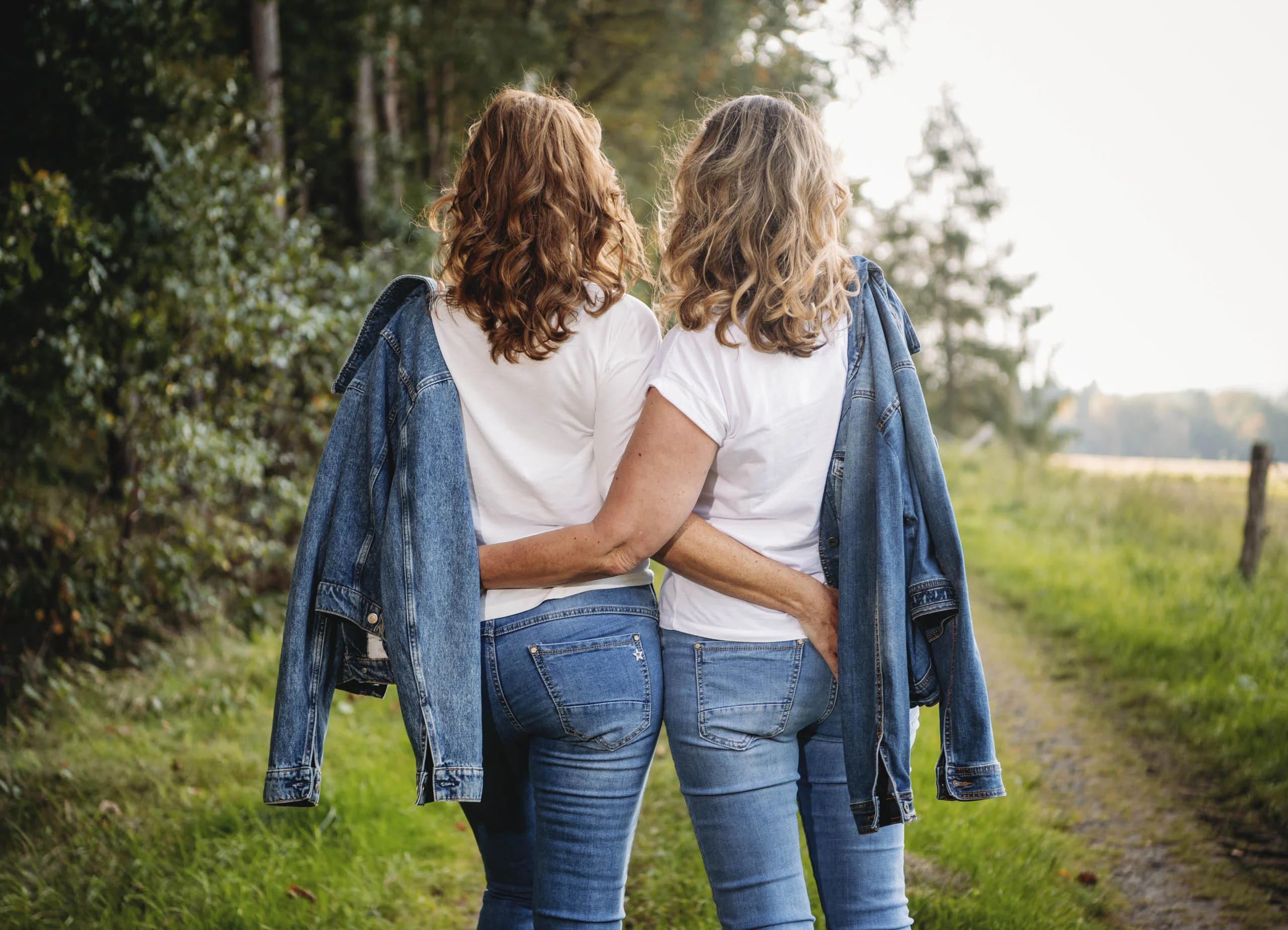 Zwei Freundinnen mit Jeansjacken über der Schulter gehen Arm in Arm einen Feldweg entlang