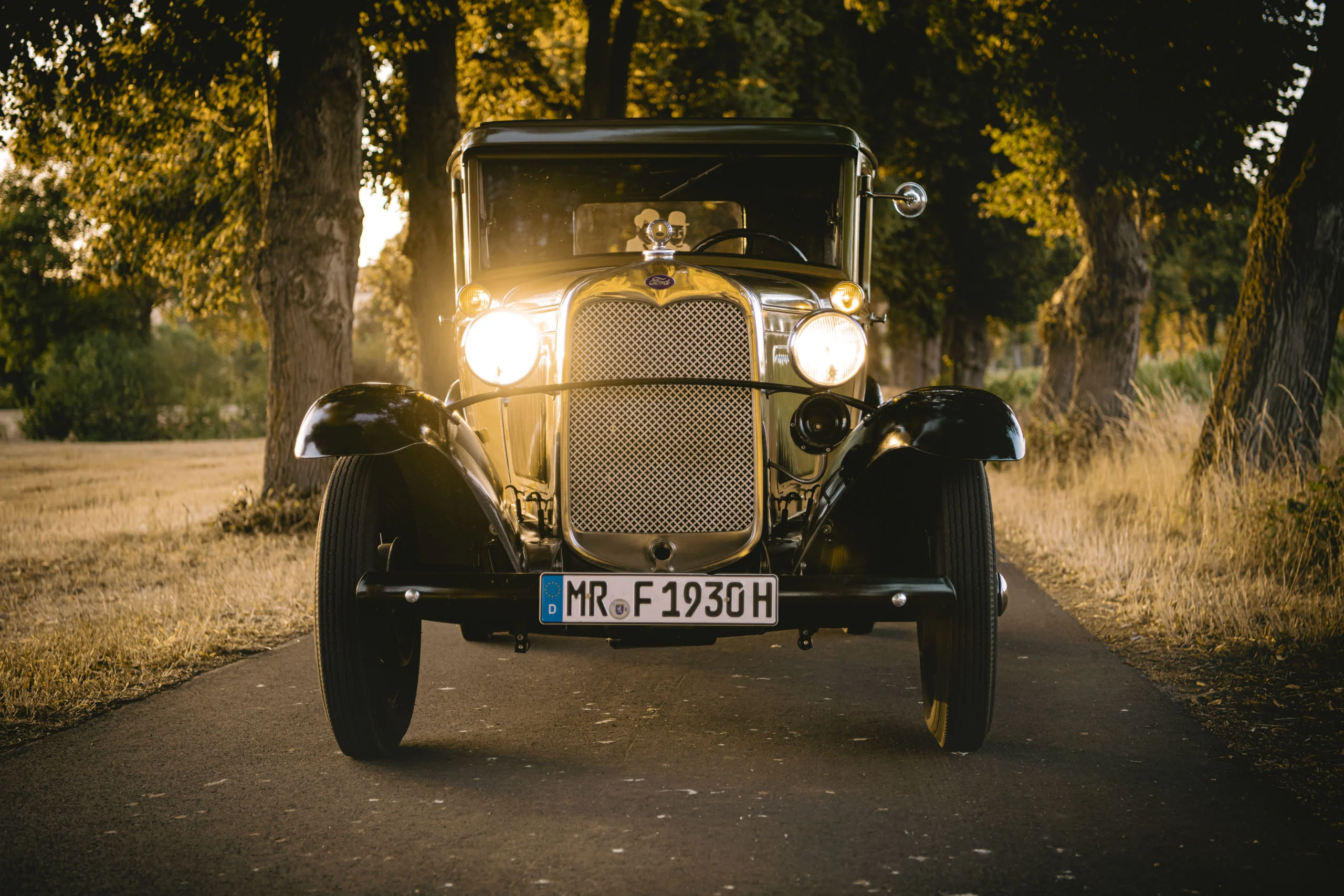 Oldtimer frontal mit leuchtenden Scheinwerfern auf einer Allee im Abendlicht