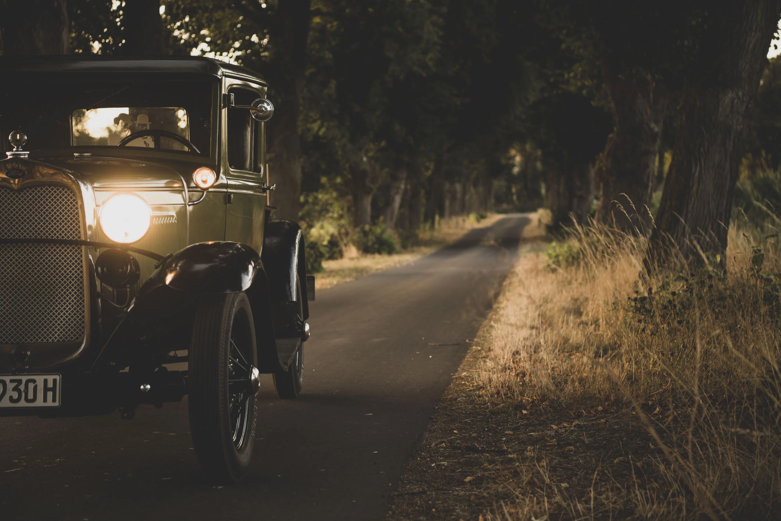 Oldtimer mit eingeschaltetem Scheinwerfer auf einem Weg durch eine Allee