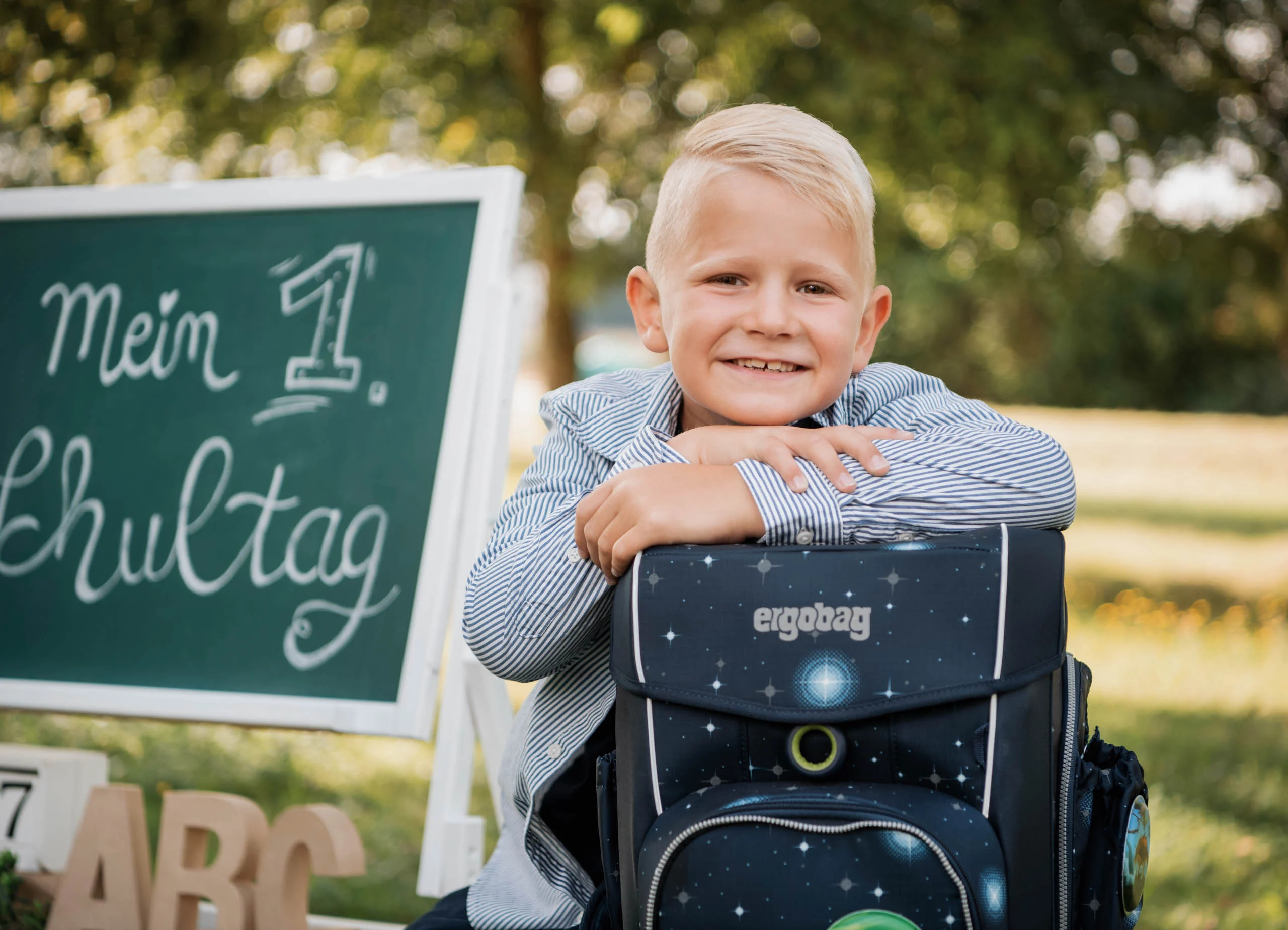 Einschulungsfotos Aktion 05 | Peter Lauritis Photography Lachender Junge stützt die Arme auf seinen Schulranzen vor der Tafel „Mein 1. Schultag“