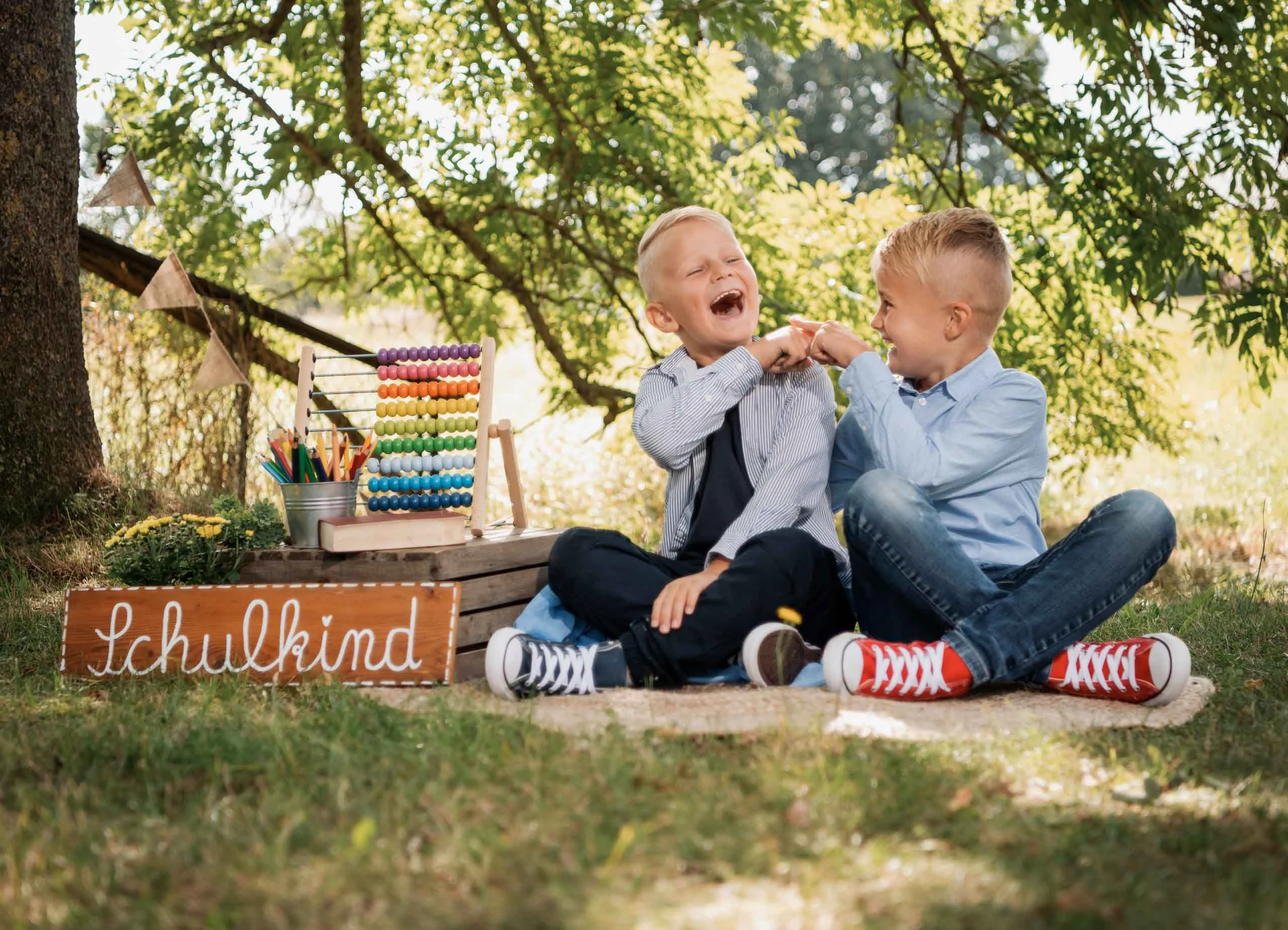 Einschulungsfotos Aktion 07 | Peter Lauritis Photography Zwei lachende Jungen sitzen im Schneidersitz vor einem Holzschild „Schulkind“ und einem bunten Rechenschieber