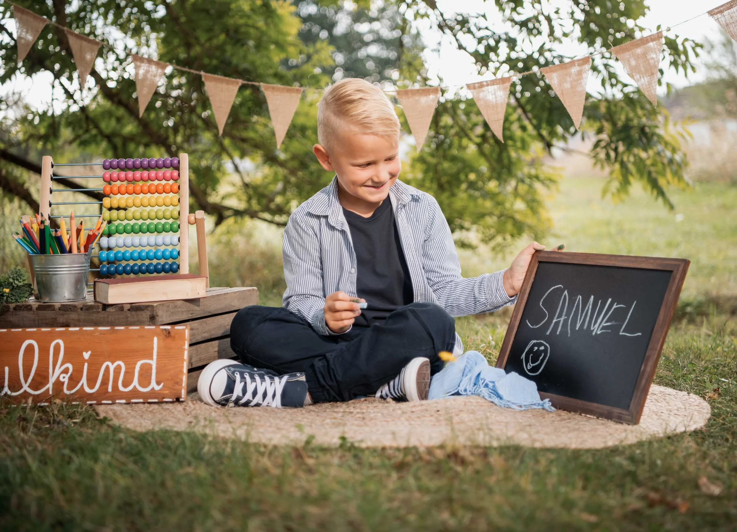 Einschulungsfotos Aktion 08 | Peter Lauritis Photography Junge sitzt im Grünen und hält eine kleine Tafel mit seinem Namen, daneben Rechenschieber und Wimpelkette