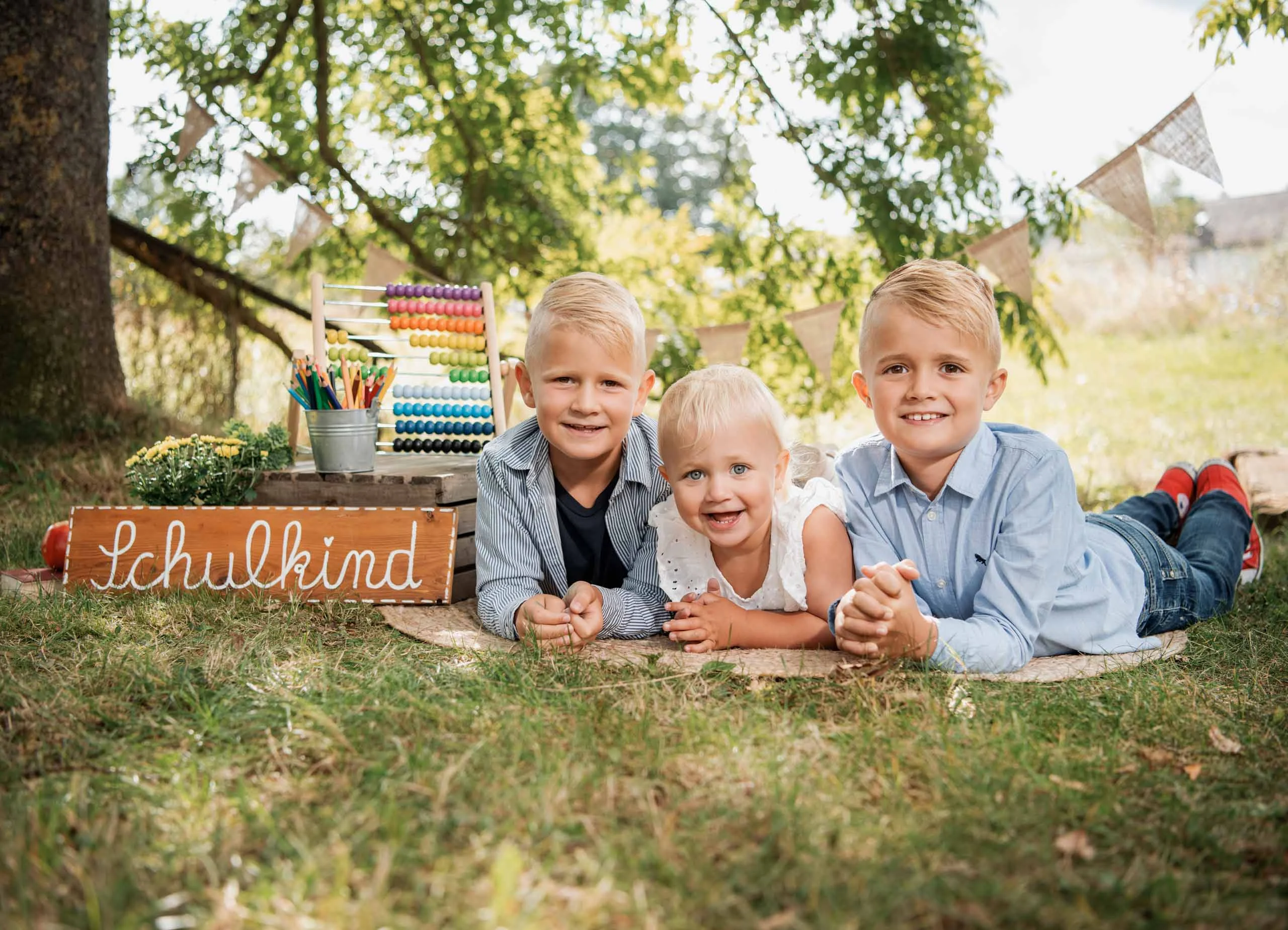 Einschulungsfotos Aktion 10 | Peter Lauritis Photography Drei Kinder liegen nebeneinander auf einer Decke im Grünen vor dem Schild „Schulkind“