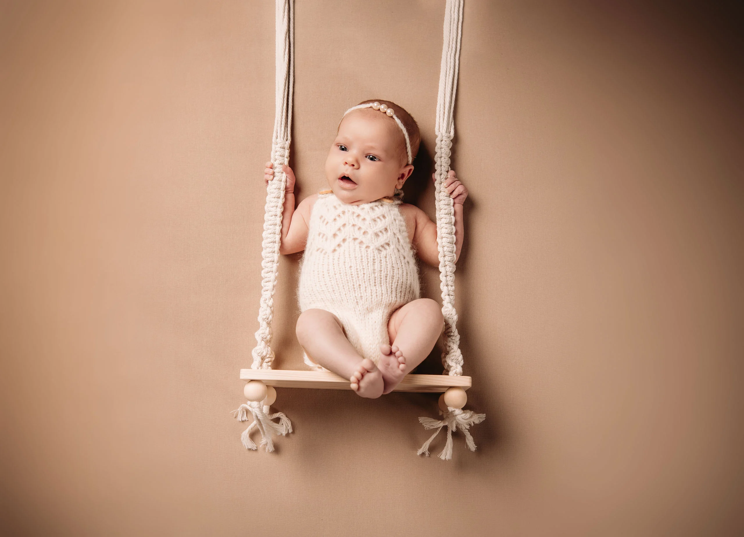 Baby im weißen Strickbody sitzt auf einer kleinen Makramee-Schaukel vor beigem Hintergrund