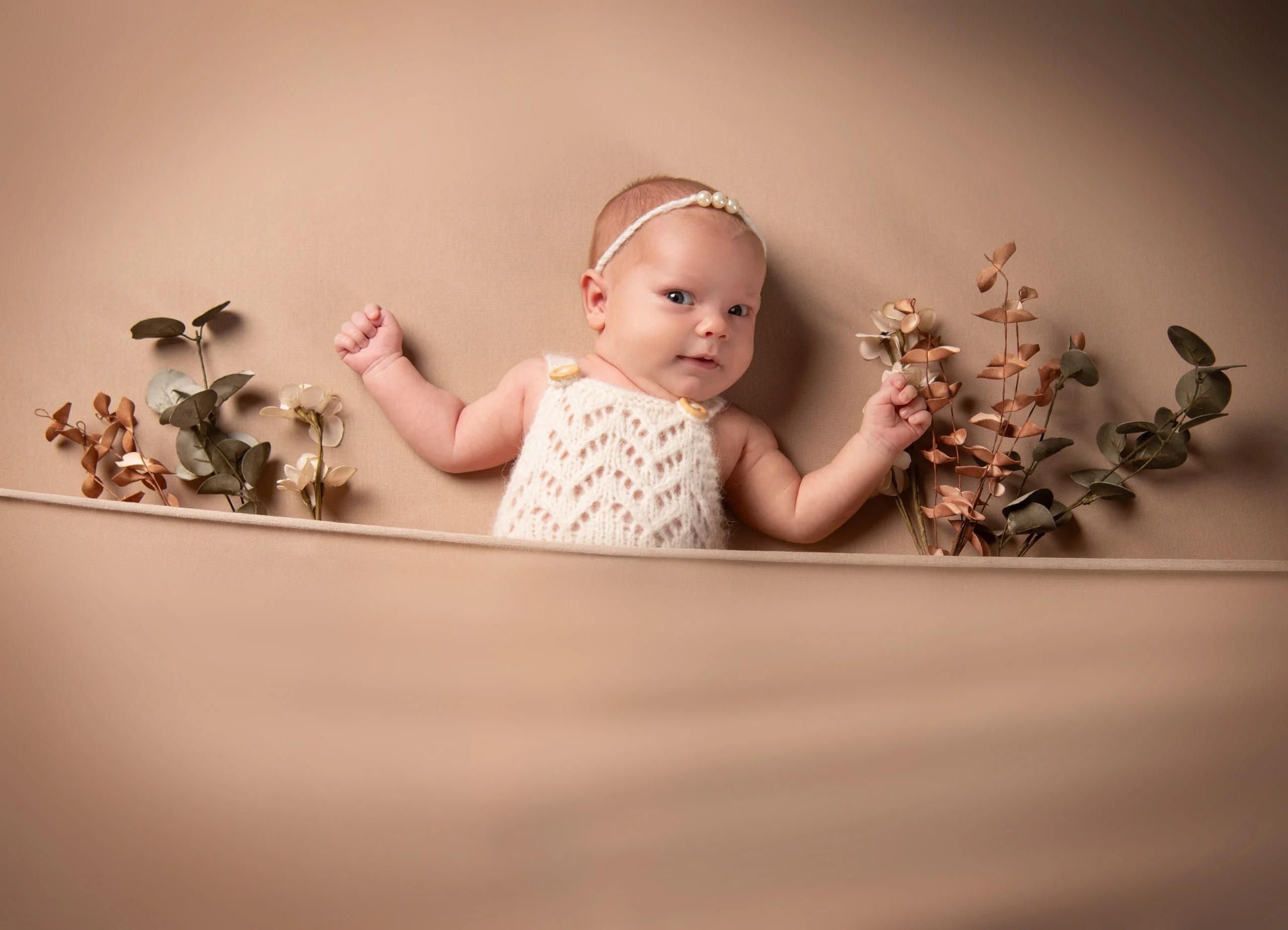 Baby- und Newbornfotos Produktbild 03 | Peter Lauritis Photography Baby im Strickbody liegt zwischen Trockenblumen auf beigem Stoff und schaut in die Kamera