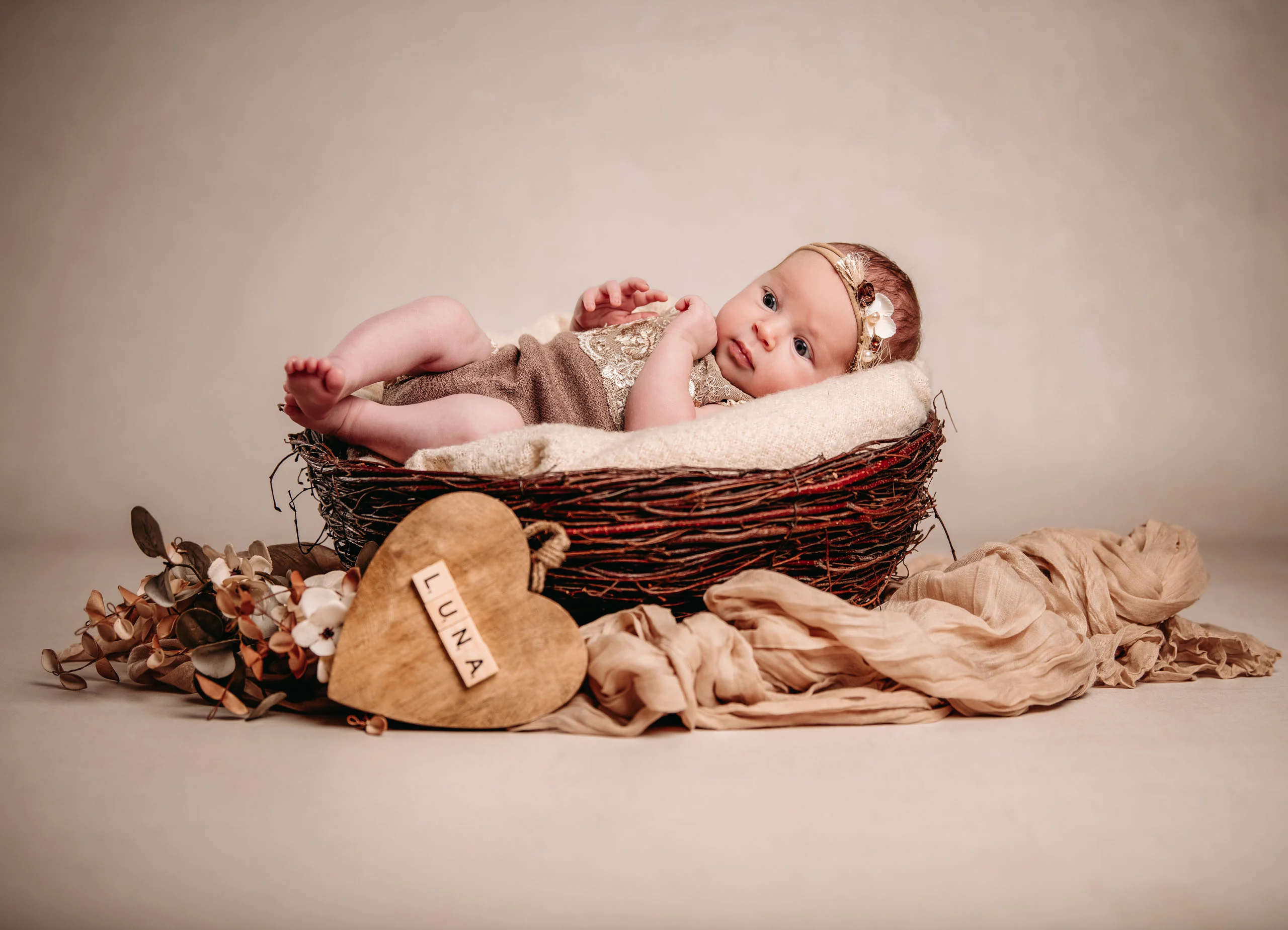 Baby- und Newbornfotos Produktbild 05 | Peter Lauritis Photography Baby mit Blumen-Haarband liegt in einem Nest-Körbchen, davor ein Holzherz mit Namen