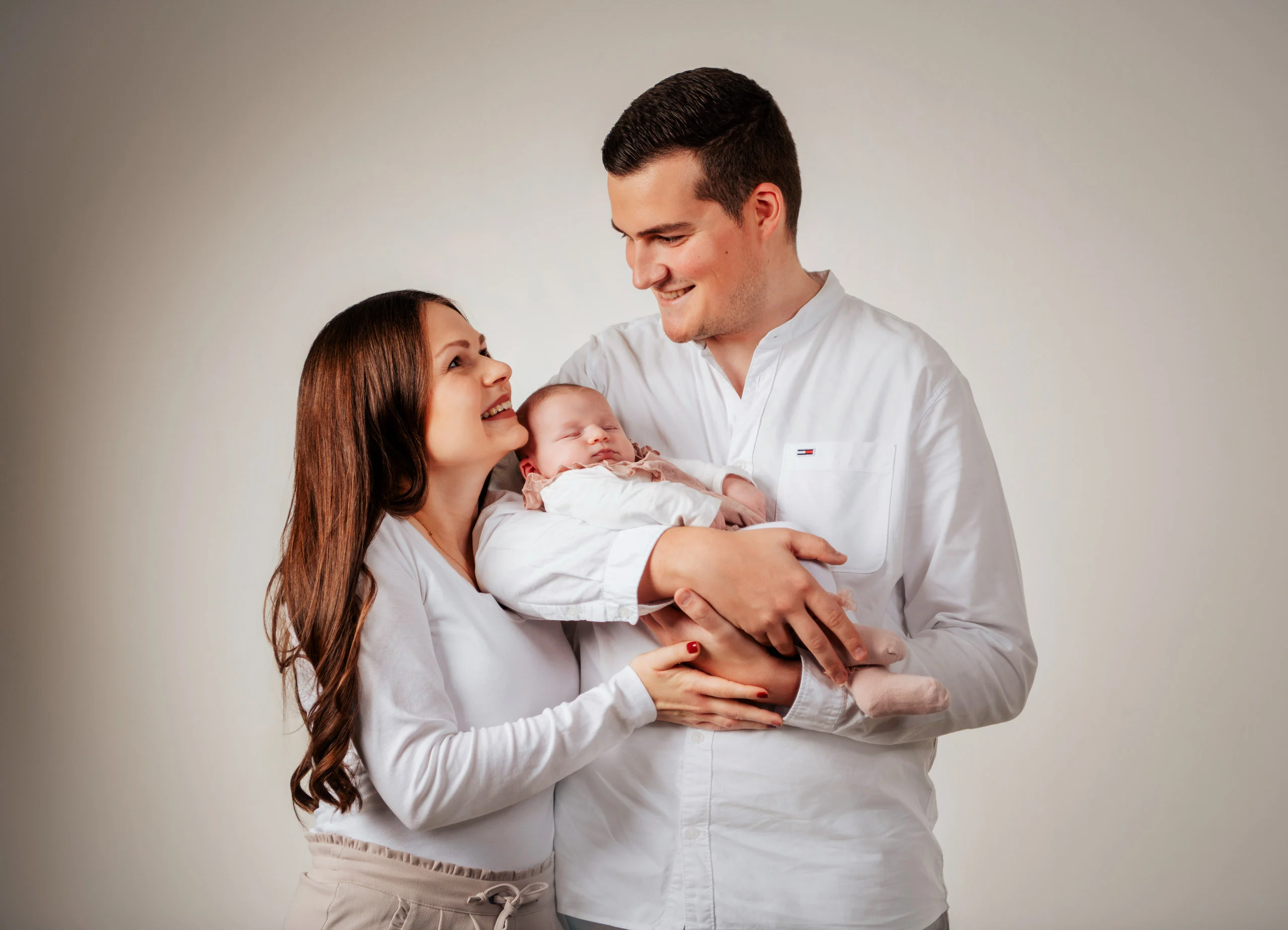 Baby- und Newbornfotos Produktbild 07 | Peter Lauritis Photography Eltern in Weiß halten ihr Neugeborenes im Arm und lächeln es an