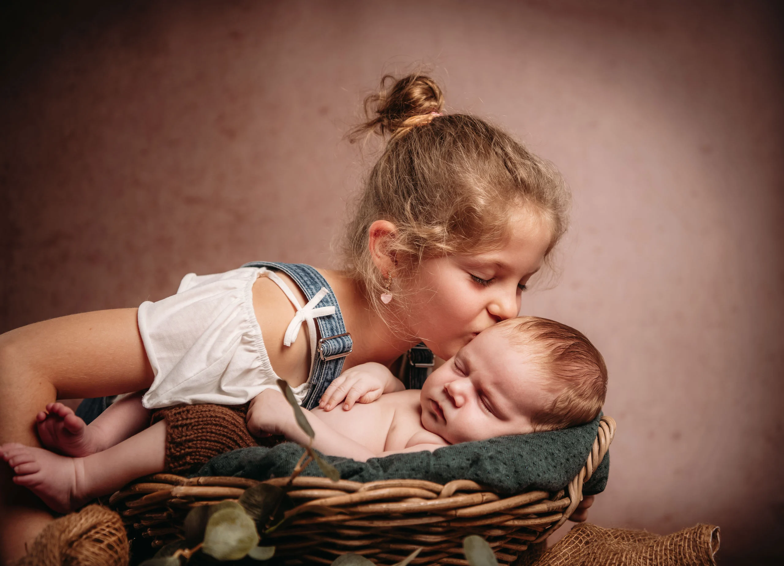 Baby- und Newbornfotos Produktbild 10 | Peter Lauritis Photography Mädchen mit Haarknoten küsst ein schlafendes Neugeborenes im Weidenkorb