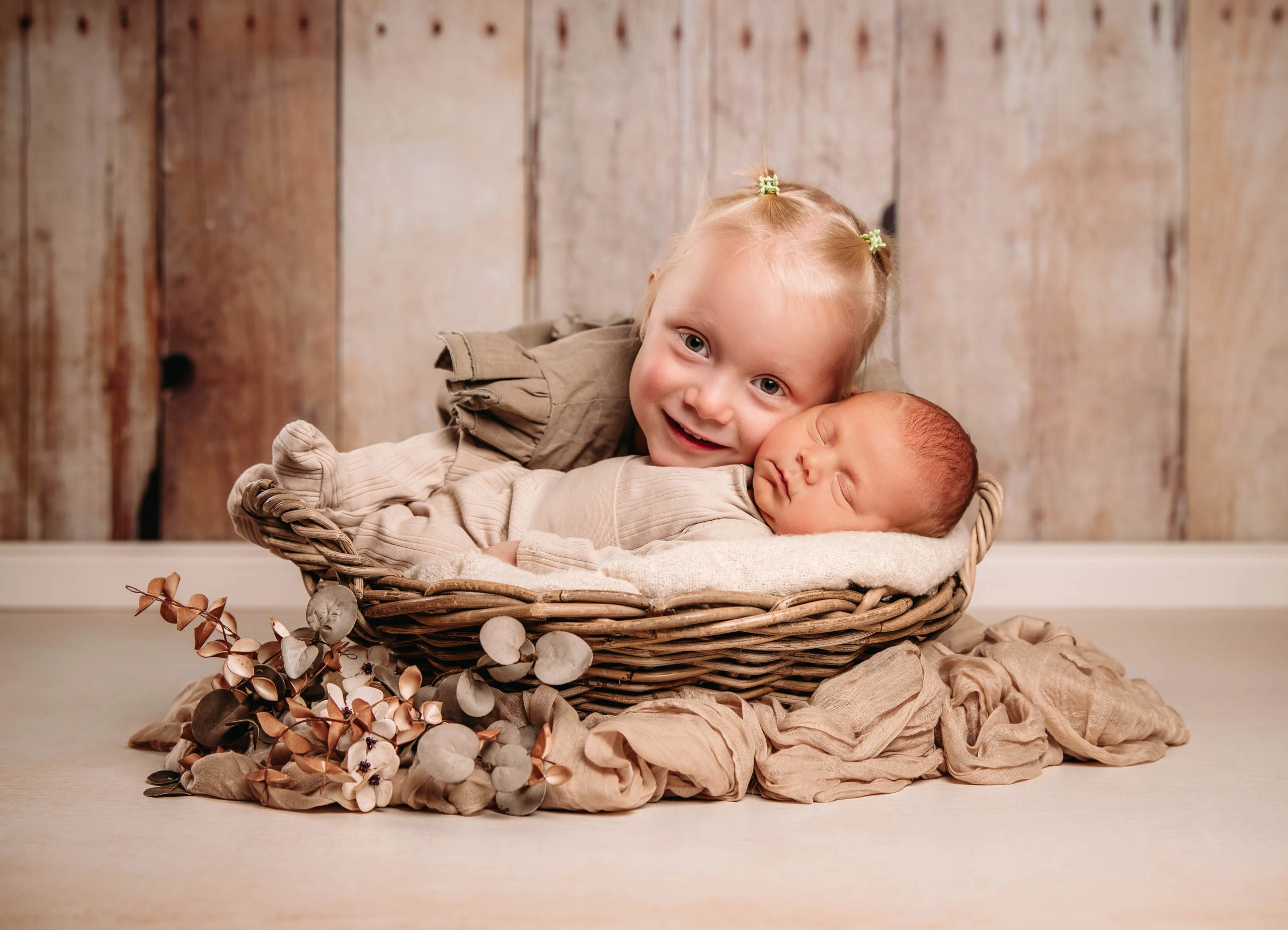 Baby- und Newbornfotos Produktbild 12 | Peter Lauritis Photography Kleinkind lehnt lachend den Kopf an ein schlafendes Neugeborenes im Korb vor Holzwand