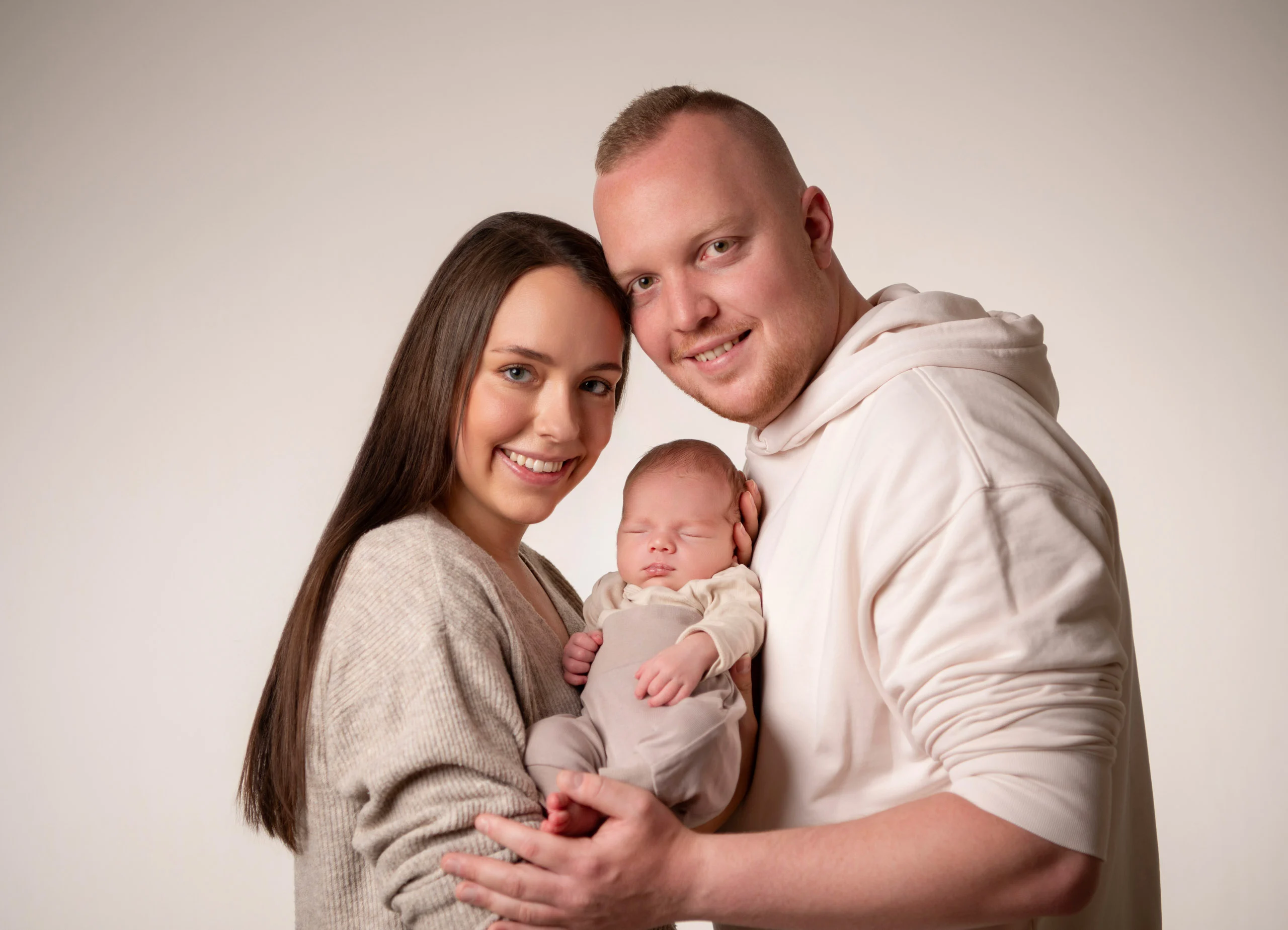 Baby- und Newbornfotos Produktbild 13 | Peter Lauritis Photography Lächelndes Paar hält sein Neugeborenes vor hellem Hintergrund