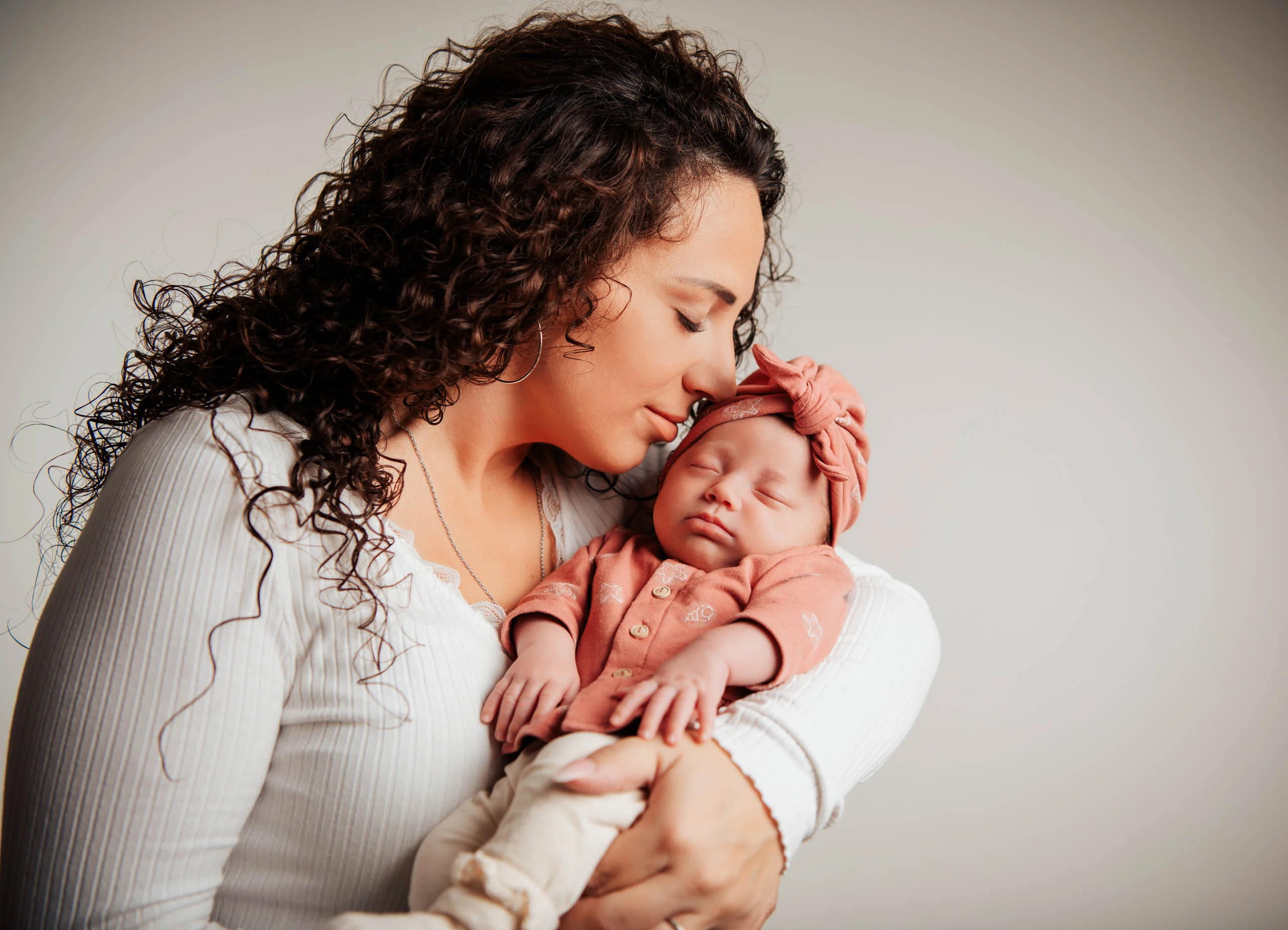 Baby- und Newbornfotos Produktbild 16 | Peter Lauritis Photography Frau mit dunklen Locken drückt ihr schlafendes Baby mit rosa Haarband an die Wange