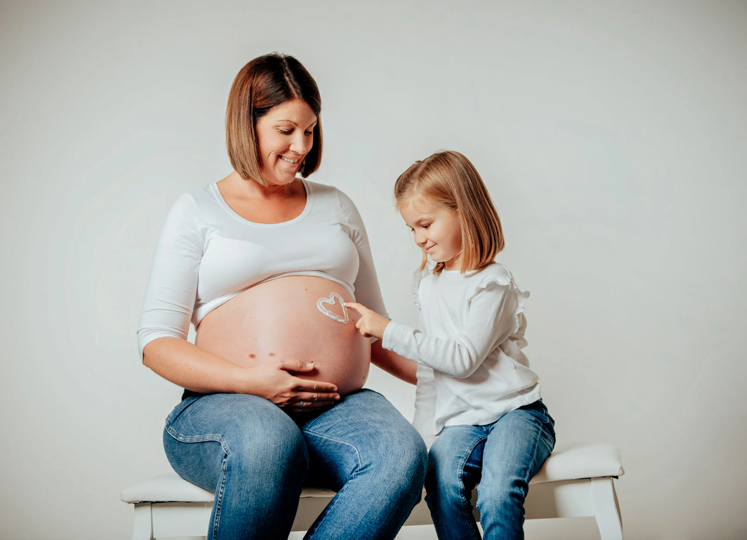 Babybauch Komplettpaket Produktbild 03 | Peter Lauritis Photography Kleines Mädchen zeigt mit einem Herz aus Fingern auf den Babybauch der sitzenden Schwangeren