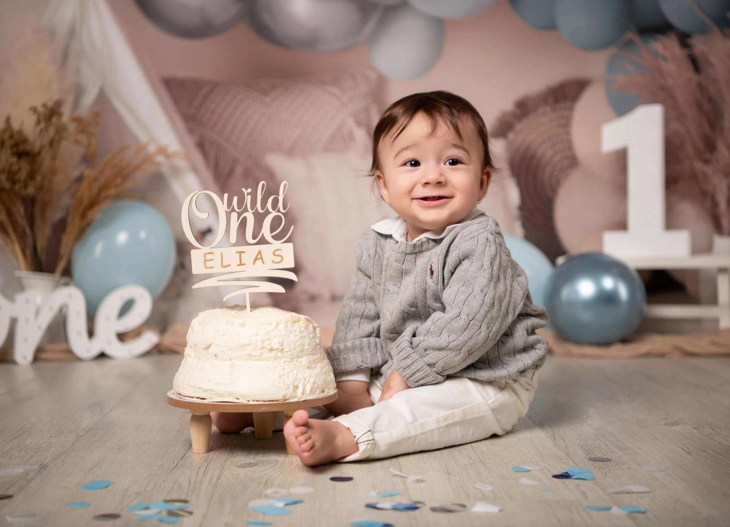 Lächelnder kleiner Junge im grauen Pullover sitzt neben einer weißen Torte mit „Wild One“-Aufsatz