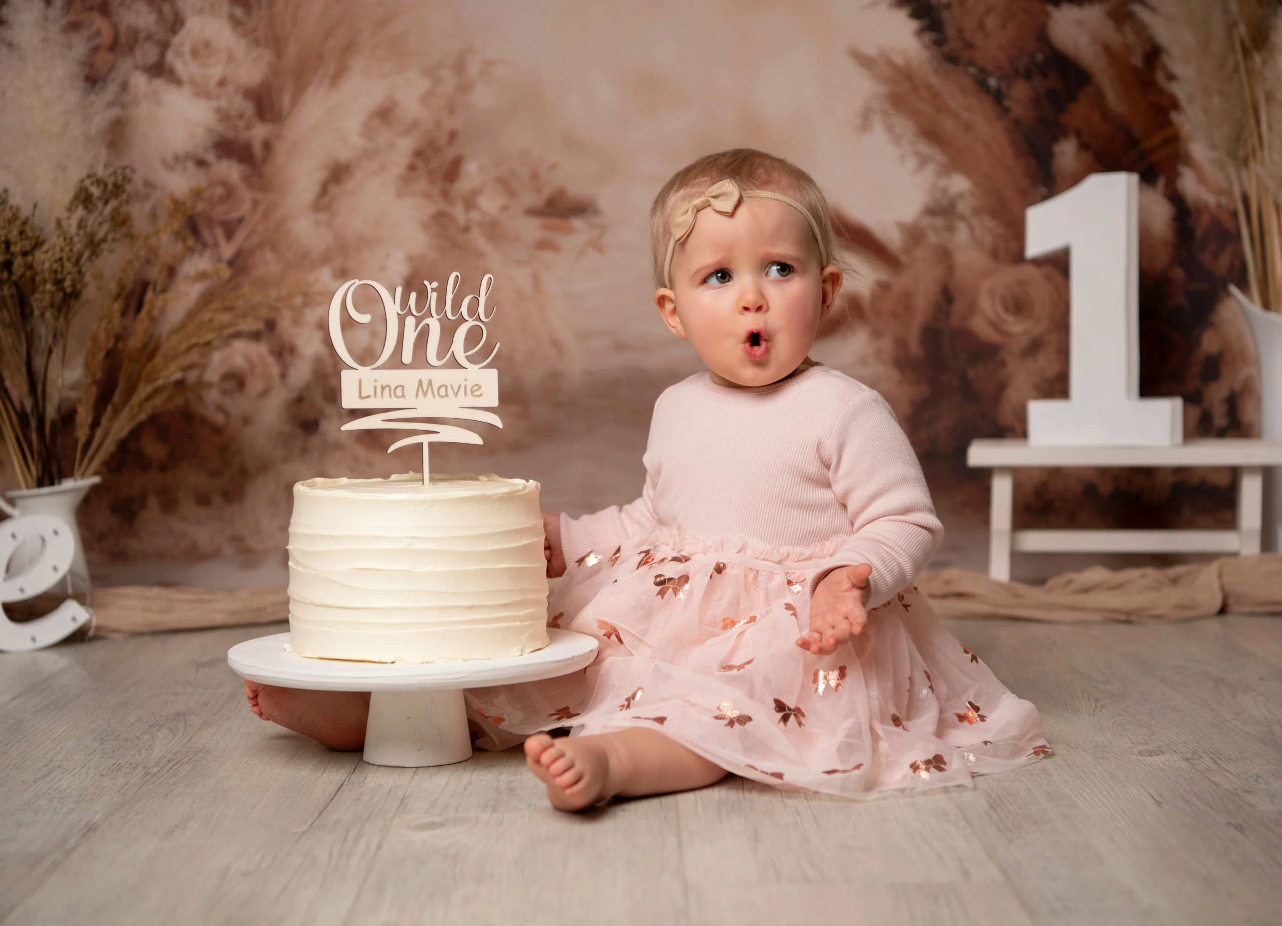 Cake Smash Produktbild 03 | Peter Lauritis Photography Staunendes Mädchen im rosa Kleid sitzt neben einer Geburtstagstorte vor Pampasgras-Kulisse