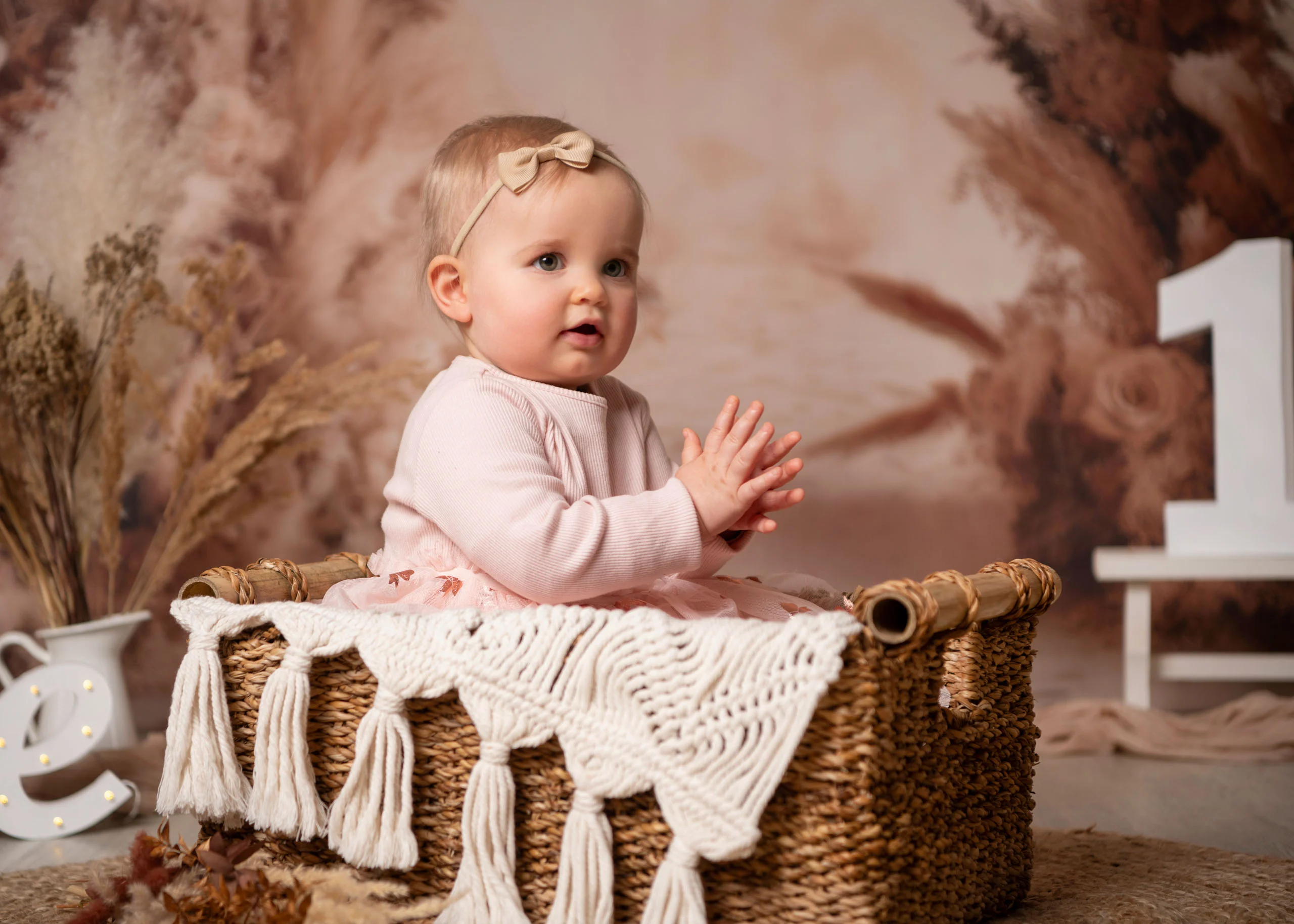 Cake Smash Produktbild 04 | Peter Lauritis Photography Kleines Mädchen mit Haarschleife sitzt in einem Korb mit Makramee-Decke vor Pampasgras-Kulisse