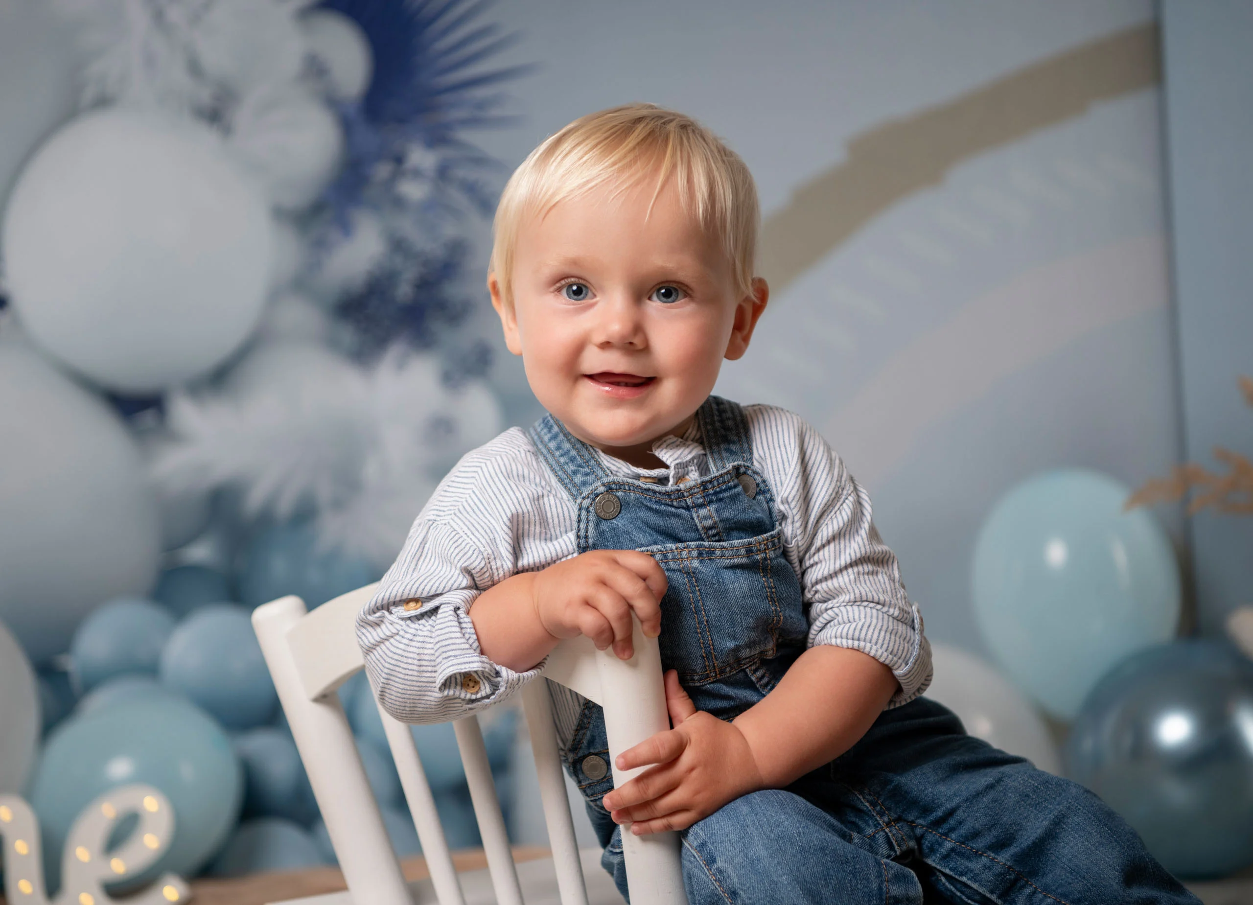 Cake Smash Produktbild 05 | Peter Lauritis Photography Lächelnder blonder Junge in Jeans-Latzhose sitzt auf einem weißen Stühlchen vor blauen Luftballons