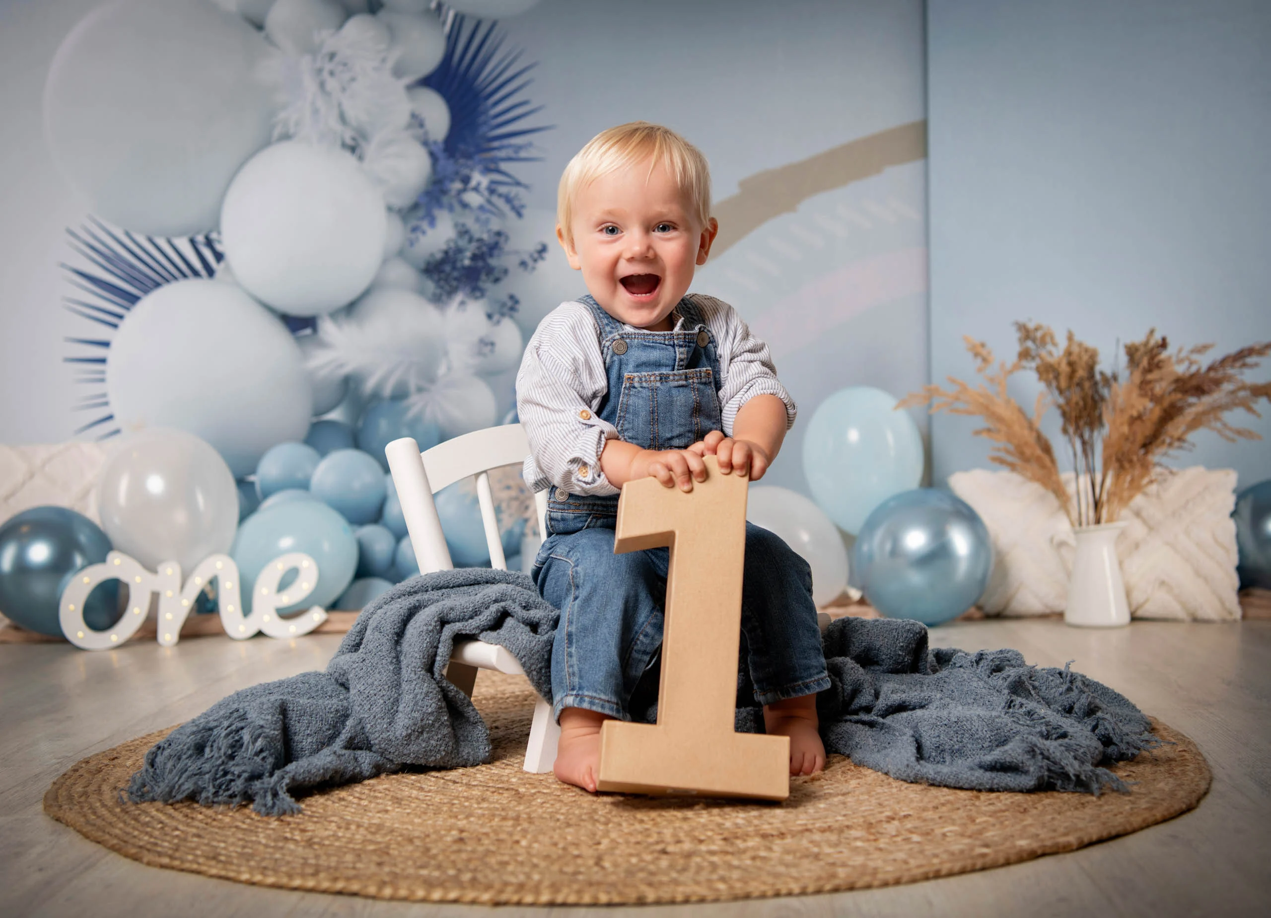Cake Smash Produktbild 06 | Peter Lauritis Photography Lachender Junge in Latzhose hält eine große Holz-Eins, hinter ihm blaue Ballonkulisse