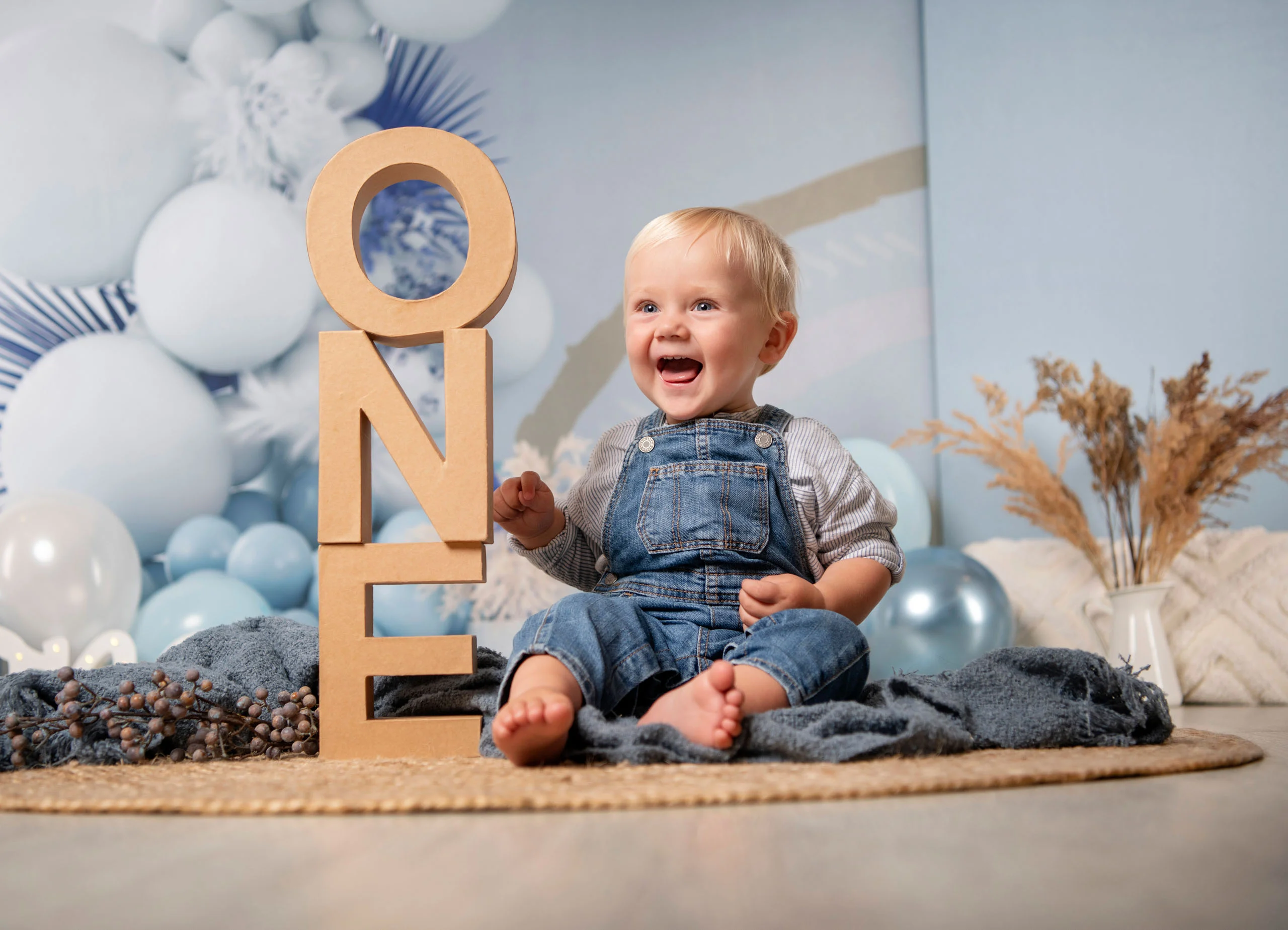 Cake Smash Produktbild 07 | Peter Lauritis Photography Lachender kleiner Junge sitzt neben den Holzbuchstaben „ONE“ vor blauen Luftballons