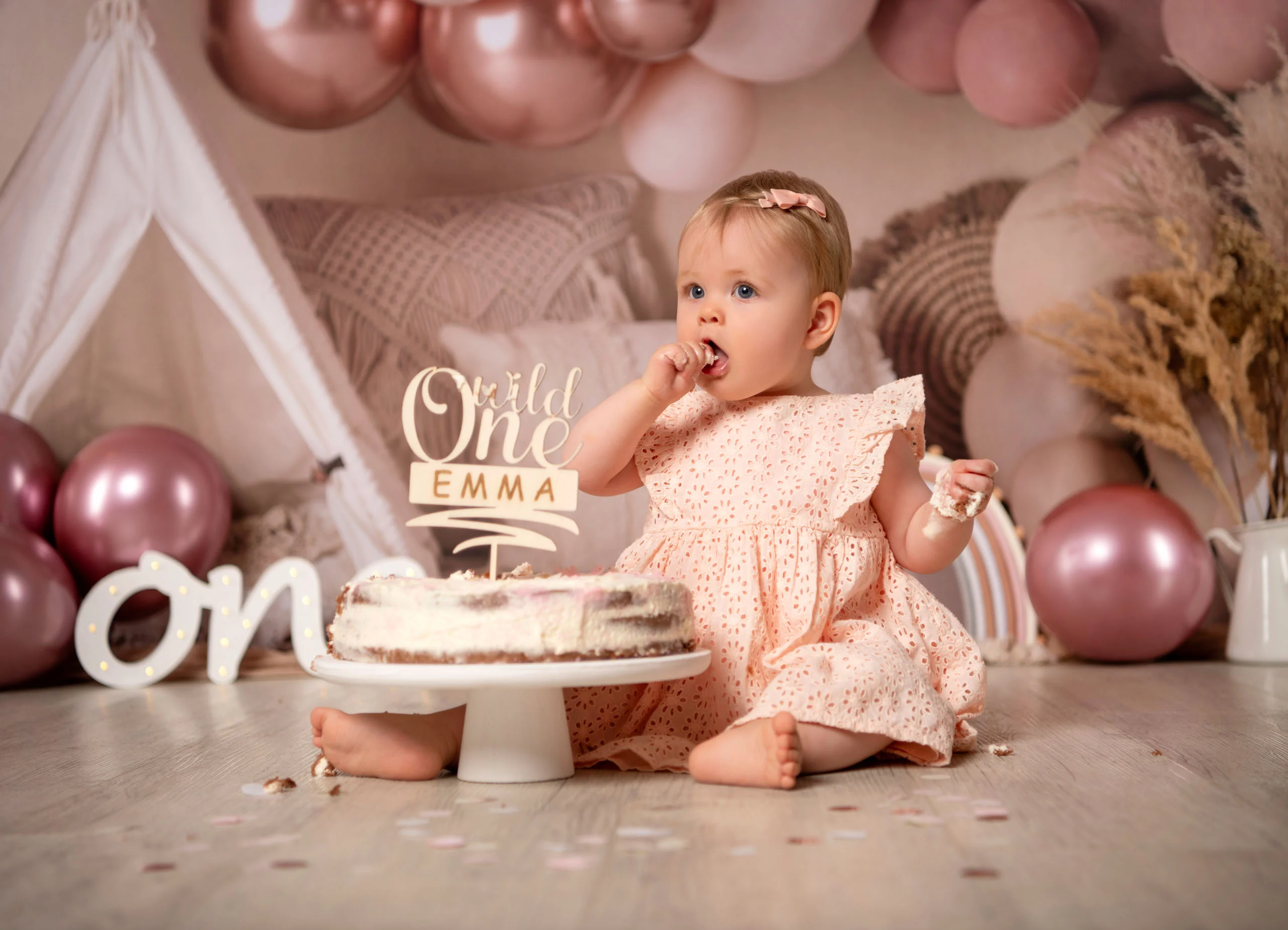 Cake Smash Produktbild 09 | Peter Lauritis Photography Kleines Mädchen schleckt Sahne von den Fingern neben der Geburtstagstorte vor rosa Luftballons