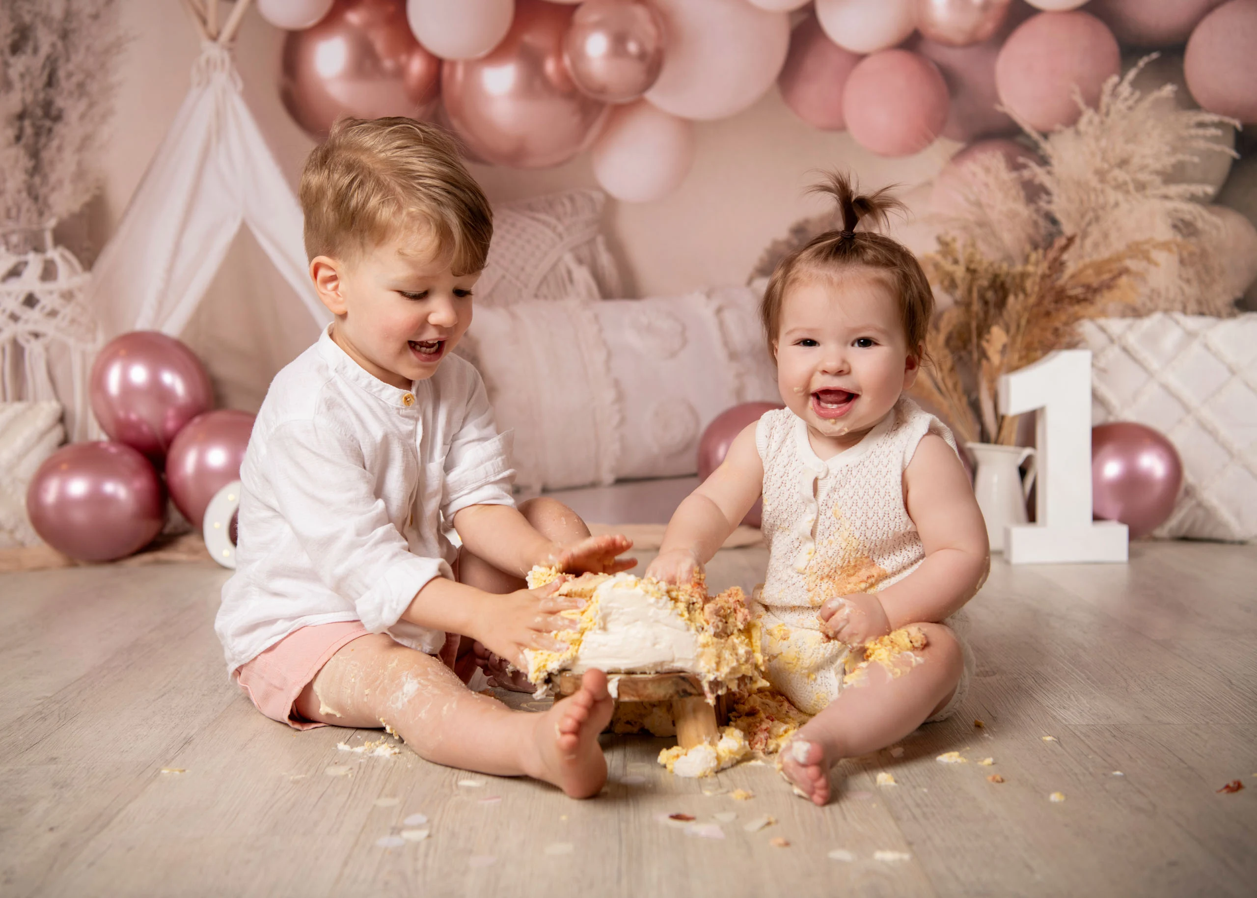 Cake Smash Produktbild 11 | Peter Lauritis Photography Zwei Kleinkinder sitzen lachend vor einer zermatschten Torte vor rosa Luftballons