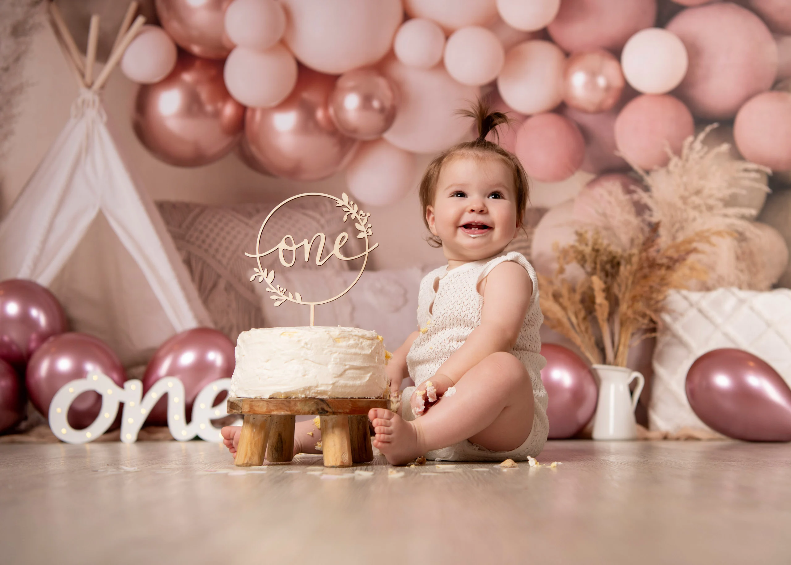 Cake Smash Produktbild 13 | Peter Lauritis Photography Kleinkind sitzt fröhlich hinter einer weißen Torte mit „one“-Aufsatz vor rosa Ballonkulisse