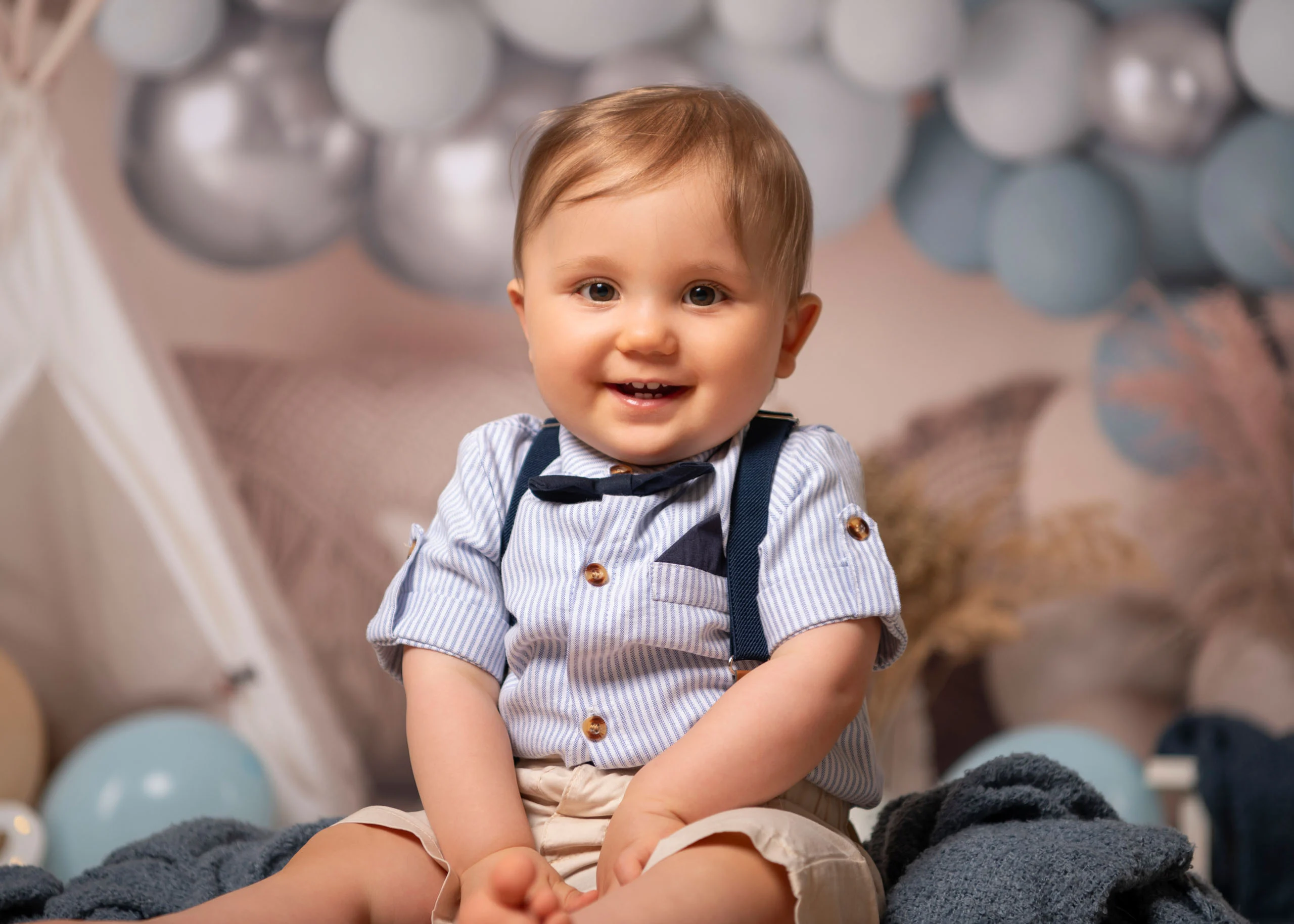 Cake Smash Produktbild 16 | Peter Lauritis Photography Lächelnder Junge mit Hosenträgern und Fliege sitzt vor grau-blauen Luftballons