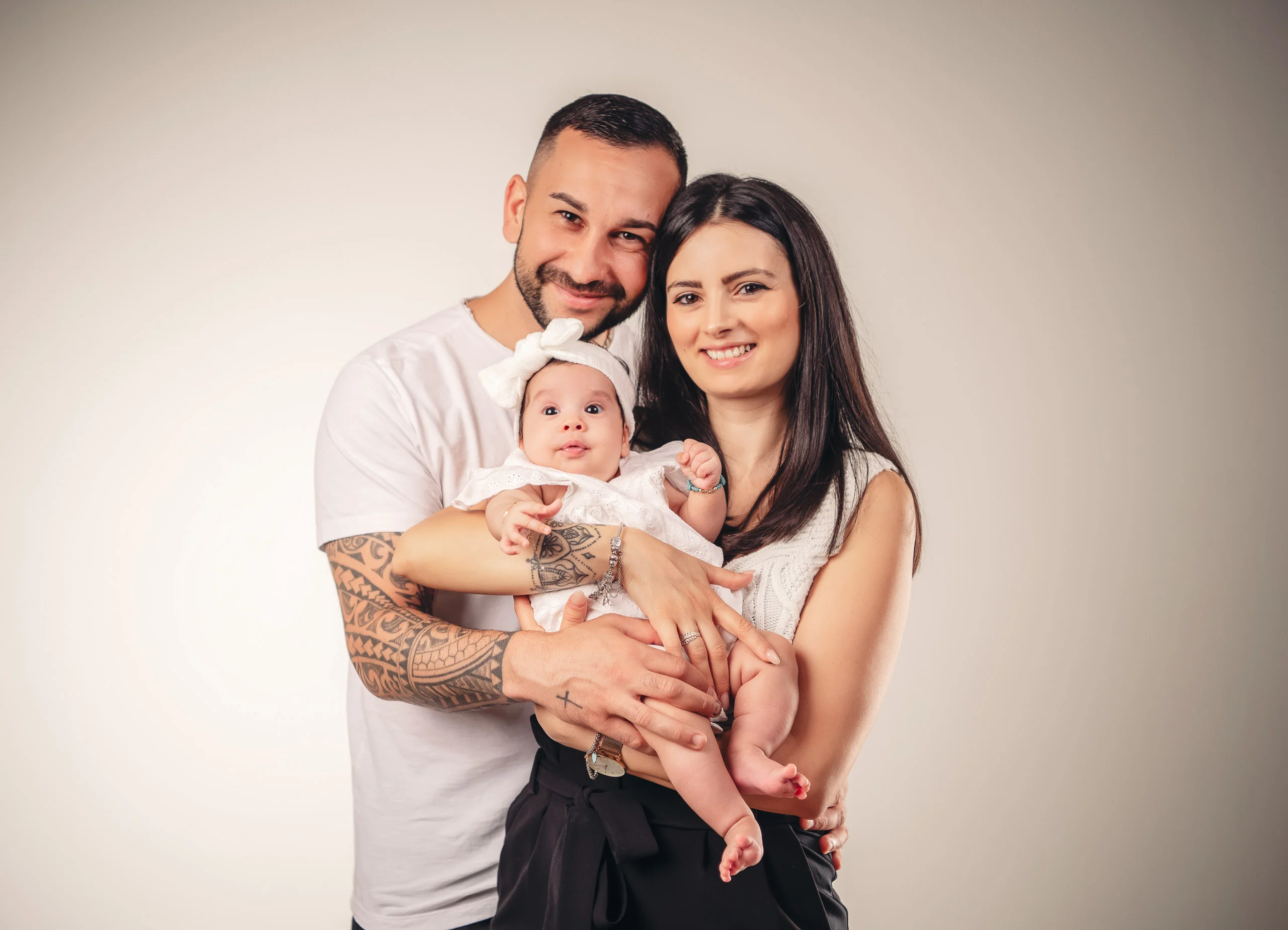 Familienfotos Produktbild 15 | Peter Lauritis Photography Paar hält ein Baby mit weißer Haarschleife zwischen sich und lächelt in die Kamera