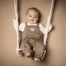 Babyfotos bei Marburg: Lachender Junge in brauner Latzhose sitzt scheinbar auf einer Makramee-Babyschaukel vor beigem Hintergrund