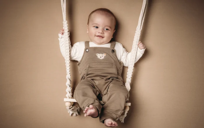 Lachendes Baby auf der Babyschaukel im Fotostudio | Peter Lauritis Photography Babyfotos bei Marburg: Lachender Junge in brauner Latzhose sitzt scheinbar auf einer Makramee-Babyschaukel vor beigem Hintergrund