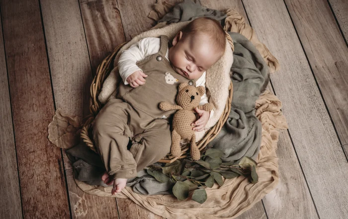 Schlafendes Baby mit Häkelteddy im Korb | Peter Lauritis Photography Baby schläft in brauner Latzhose in einem Korb auf Holzboden und kuschelt mit einem gehäkelten Teddy, daneben Eukalyptuszweige