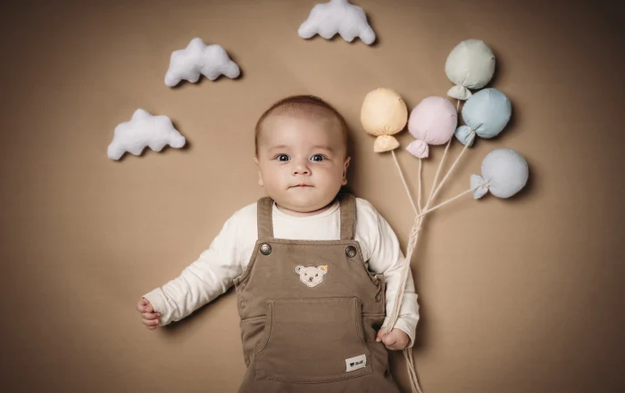 Baby mit Filzwolken und Luftballons | Peter Lauritis Photography Baby in Latzhose liegt auf beigem Hintergrund zwischen weißen Filzwolken und hält bunte Filz-Luftballons in Pastell