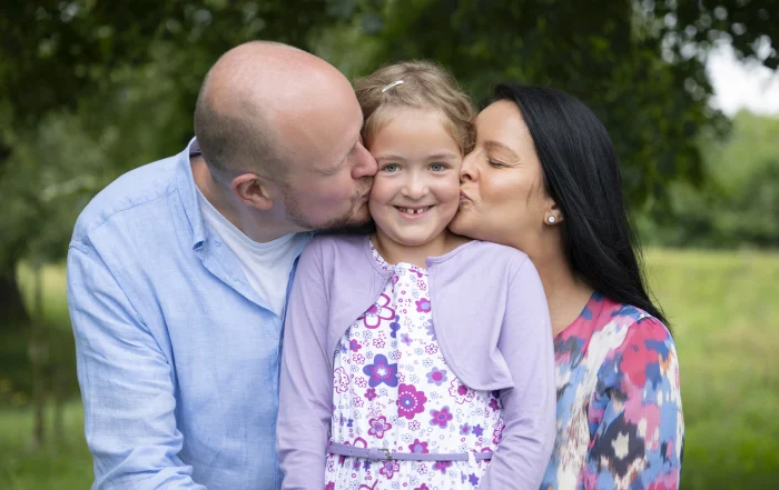 Eltern küssen ihr Schulkind | Peter Lauritis Photography Mama und Papa geben ihrer Tochter zur Einschulung gleichzeitig einen Kuss auf beide Wangen, im Hintergrund grüne Bäume