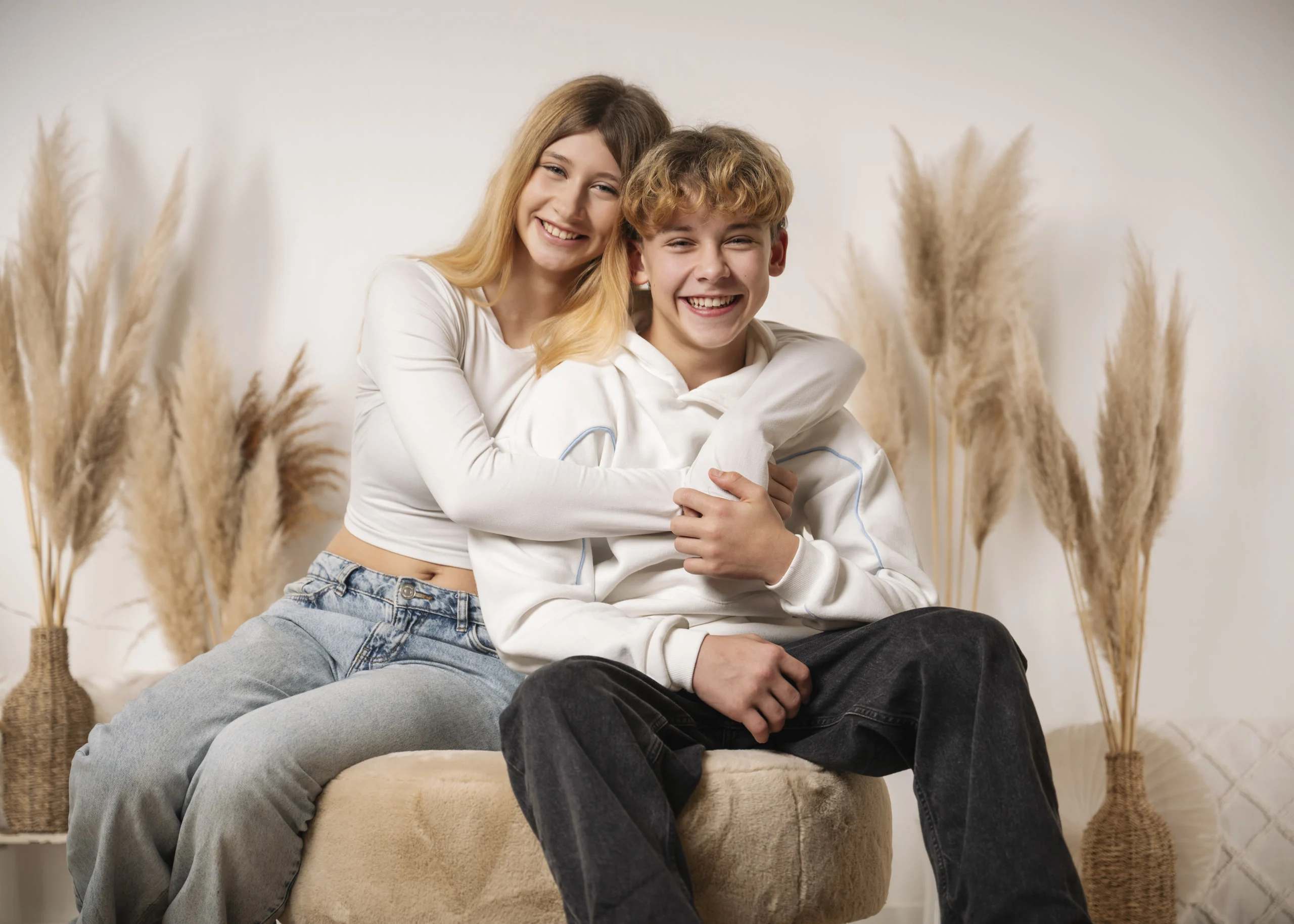 Geschwister lachen beim Fotoshooting vor Weihnachten | Peter Lauritis Photography | Peter Lauritis Photography Fotoshooting vor Weihnachten: Teenager-Schwester umarmt ihren Bruder, beide sitzen lachend auf einem Hocker vor Pampasgras