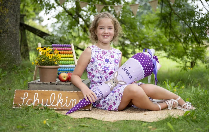 Schulkind mit Schultüte | Peter Lauritis Photography Fotoshooting zur Einschulung: Mädchen sitzt mit lila Schultüte vor einem Holzschild Schulkind, daneben ein bunter Rechenschieber