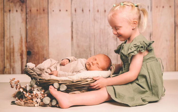 Große Schwester bewundert ihren neugeborenen Bruder im Korb | Peter Lauritis Photography Neugeborenenfotos mit Geschwisterkind: Große Schwester im grünen Kleid lächelt ihren neugeborenen Bruder im Weidenkorb an