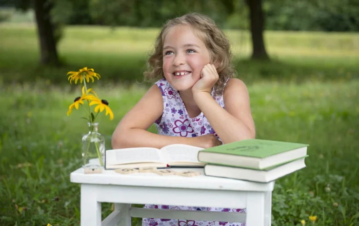 Schulkind am kleinen Schreibtisch | Peter Lauritis Photography Mädchen sitzt an einem kleinen weißen Schreibtisch mit Büchern und Blumen auf der Wiese und lacht