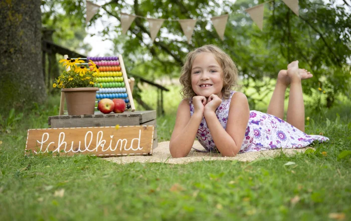 Schulkind liegt im Gras | Peter Lauritis Photography Mädchen liegt auf dem Bauch im Gras, stützt das Kinn auf die Hände und lächelt, daneben das Schild Schulkind