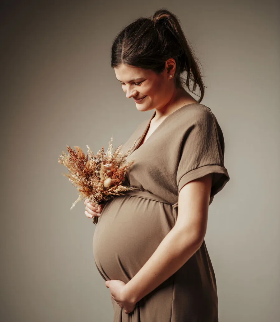 startseite babybauch fotoshooting Kachel | Peter Lauritis Photography Schwangere Frau im braunen Kleid hält lächelnd ihren Babybauch und einen Trockenblumenstrauß – Babybauchfotos im Fotostudio