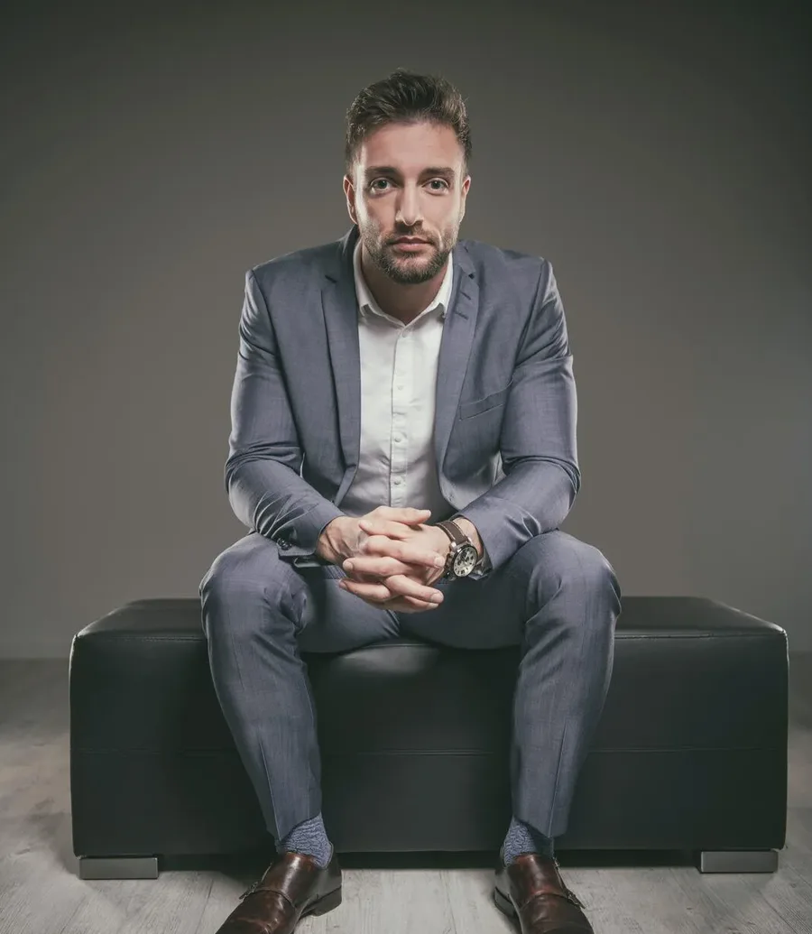 startseite business portraits Kachel | Peter Lauritis Photography Business-Porträt: Mann im grauen Anzug sitzt mit gefalteten Händen auf einer schwarzen Bank im Fotostudio
