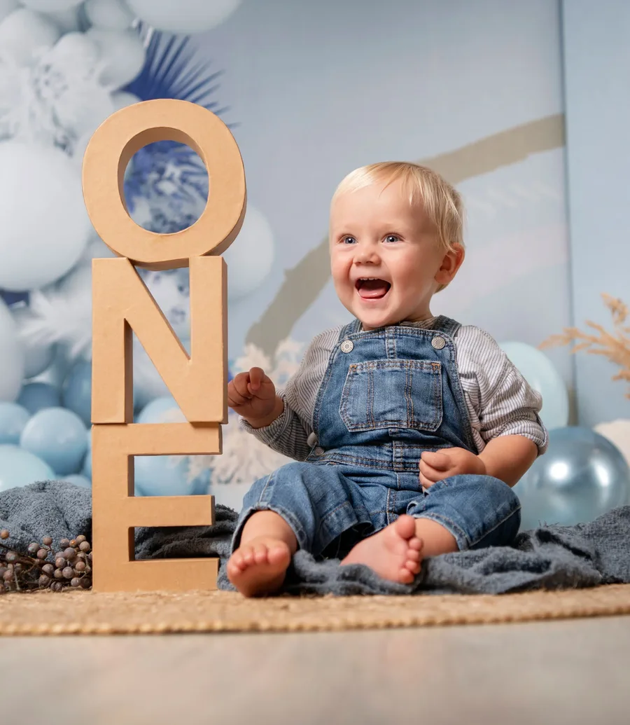 startseite cake smash fotoshooting Kachel | Peter Lauritis Photography Lachender Junge im Jeans-Latzanzug neben den Holzbuchstaben ONE beim Cake Smash Fotoshooting